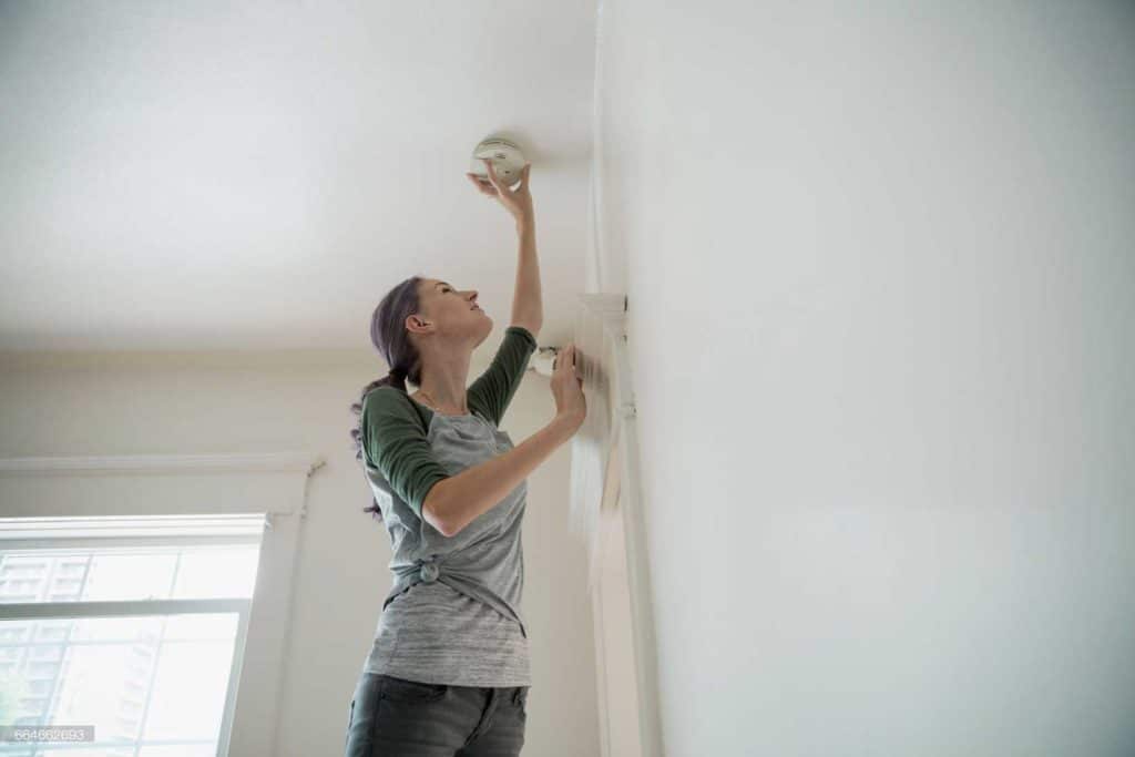 Woman standing on tiptoe to install or test a smoke detector on the ceiling.
